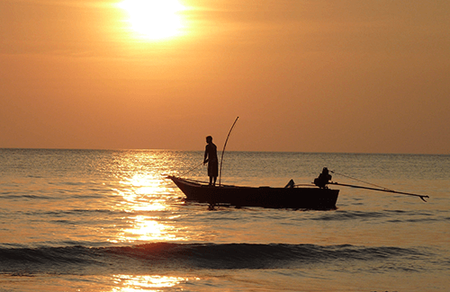 Man fishing from deck of fishing boat on the ocean as the sun sets.